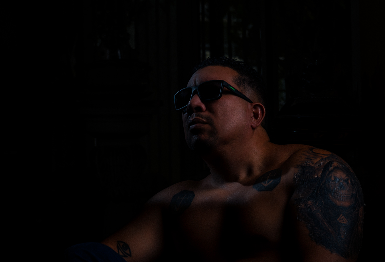 Retrato sentado con gafas oscuras, mirada elevada y tatuajes bajo una luz tenue.