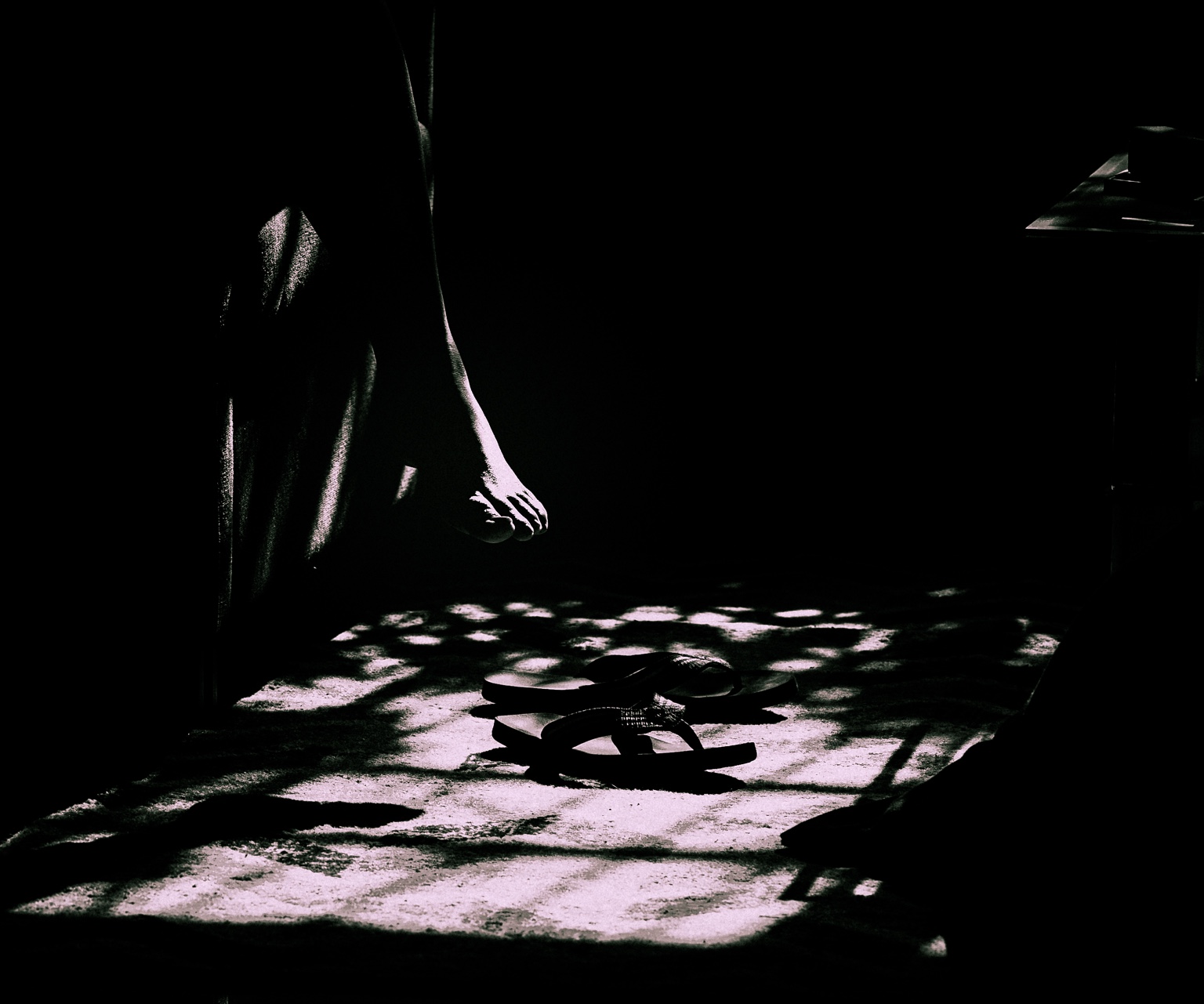 Un pie suspendido sobre unas sandalias, entre sombras profundas y manchas de luz en el suelo.