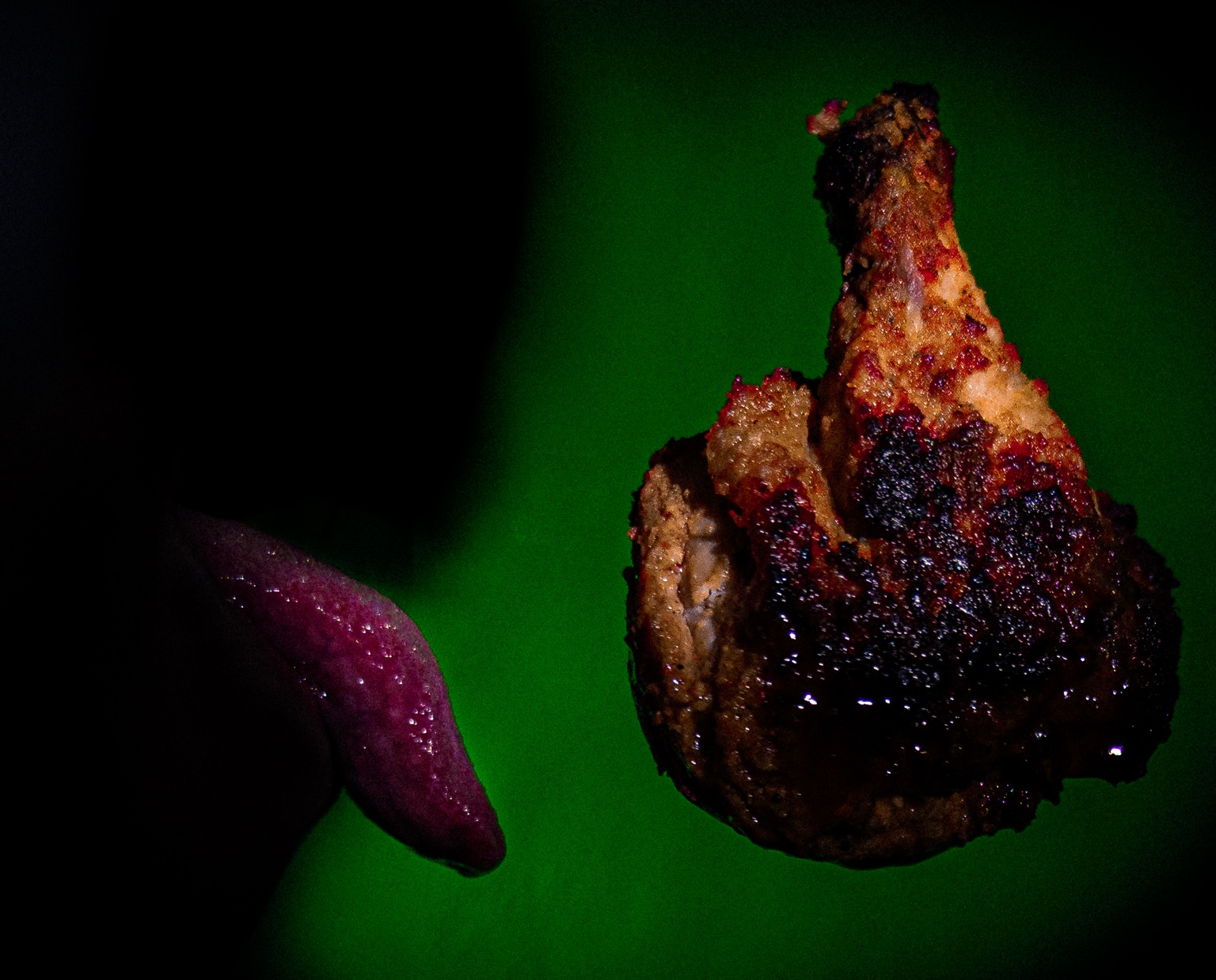 Una lengua emerge de la sombra y se acerca a una pieza de carne suspendida sobre un fondo verde.