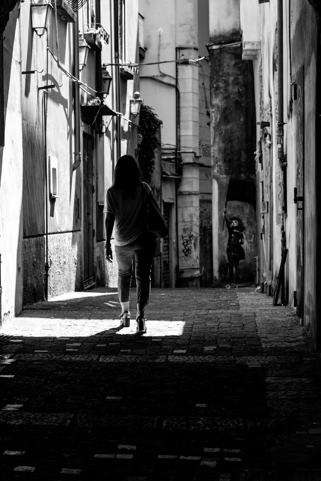 Una mujer de espaldas camina por un callejón entre franjas de luz y sombra.