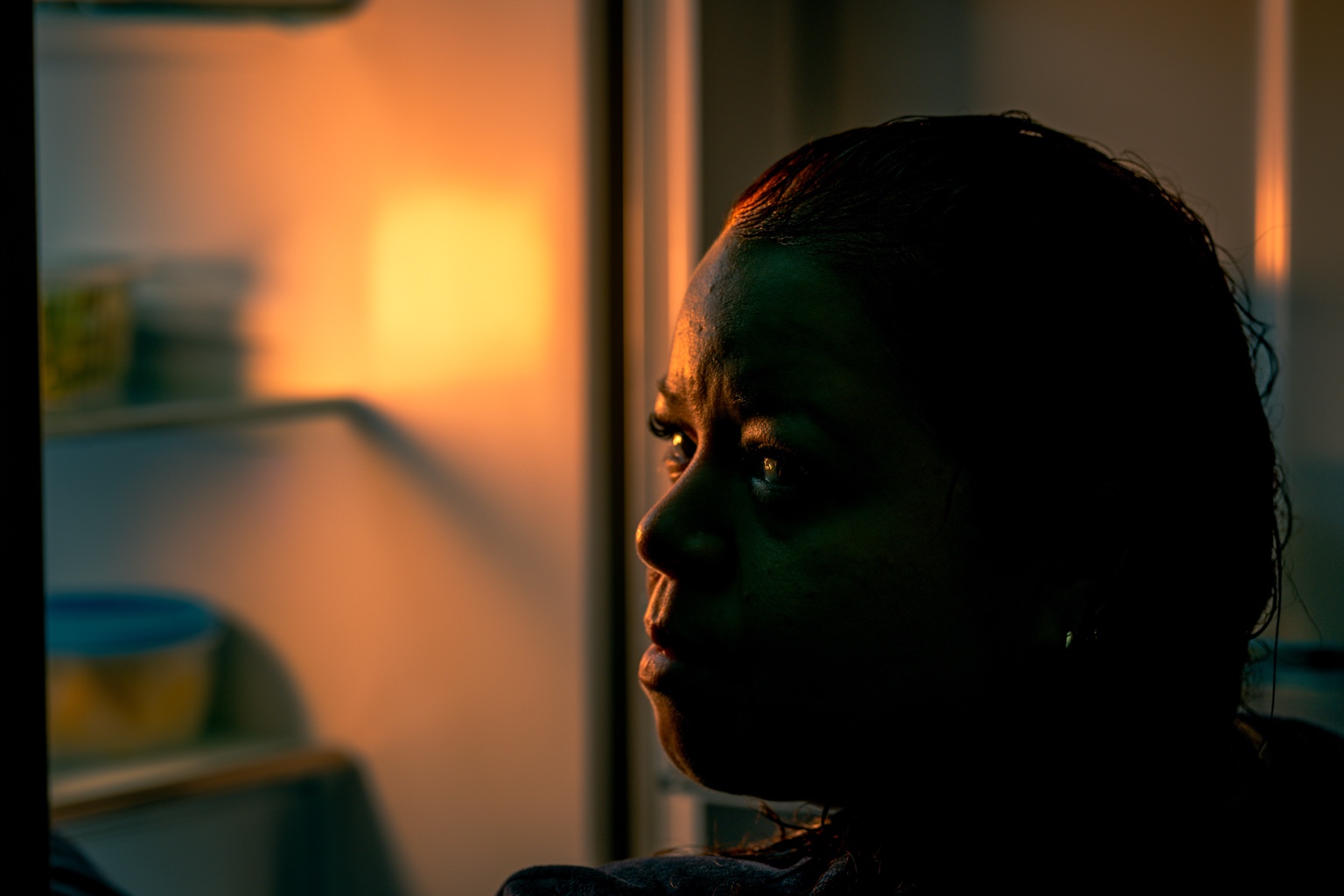 Una mujer de perfil junto a una nevera abierta, entre luz cálida y sombras frías.