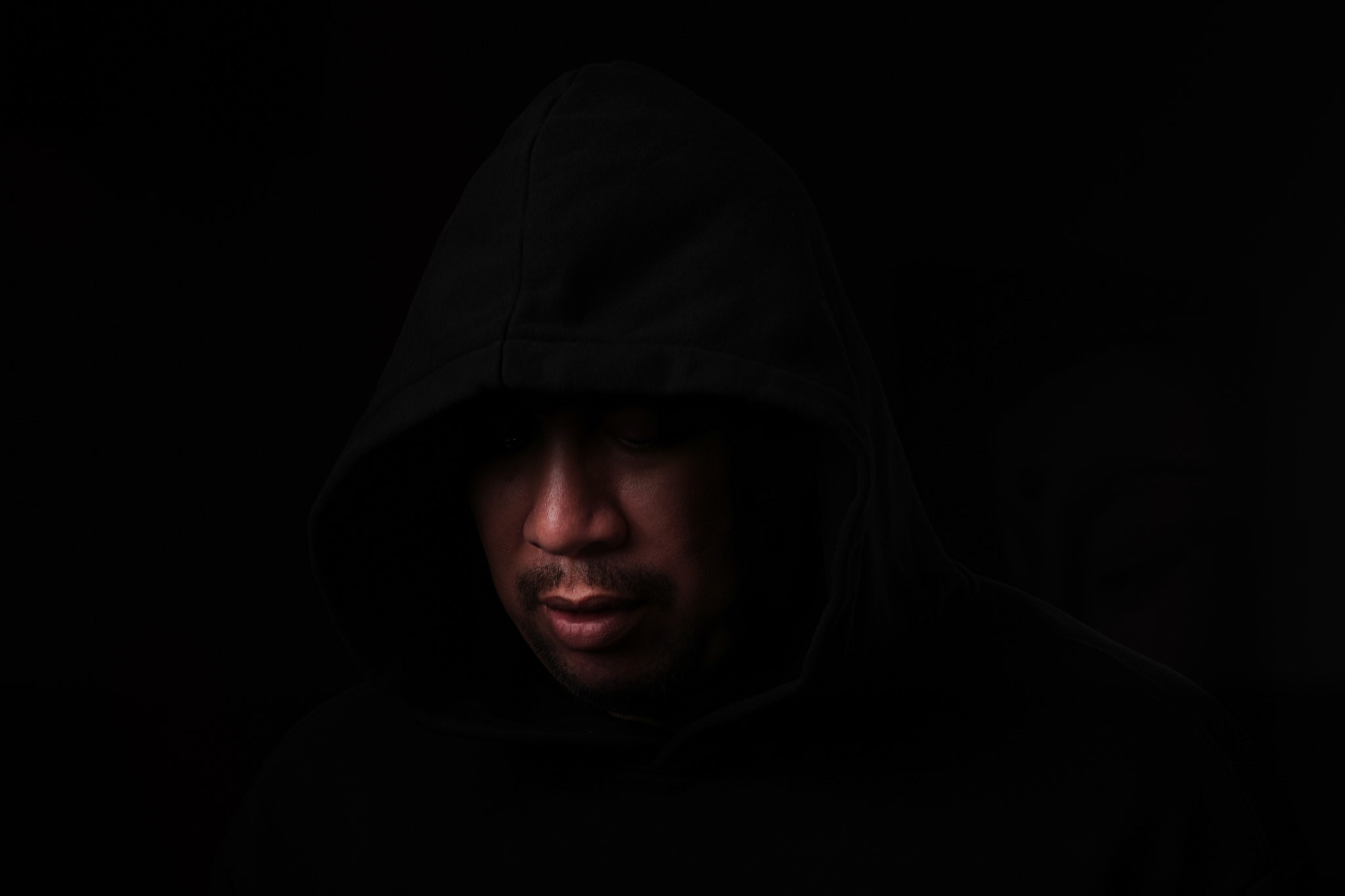 Retrato con capucha negra y la mirada hacia abajo, con el rostro parcialmente iluminado sobre fondo oscuro.