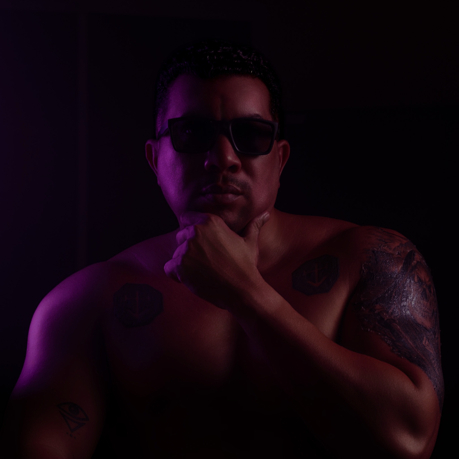 Retrato frontal con la mano bajo el mentón, gafas oscuras e iluminación violeta.