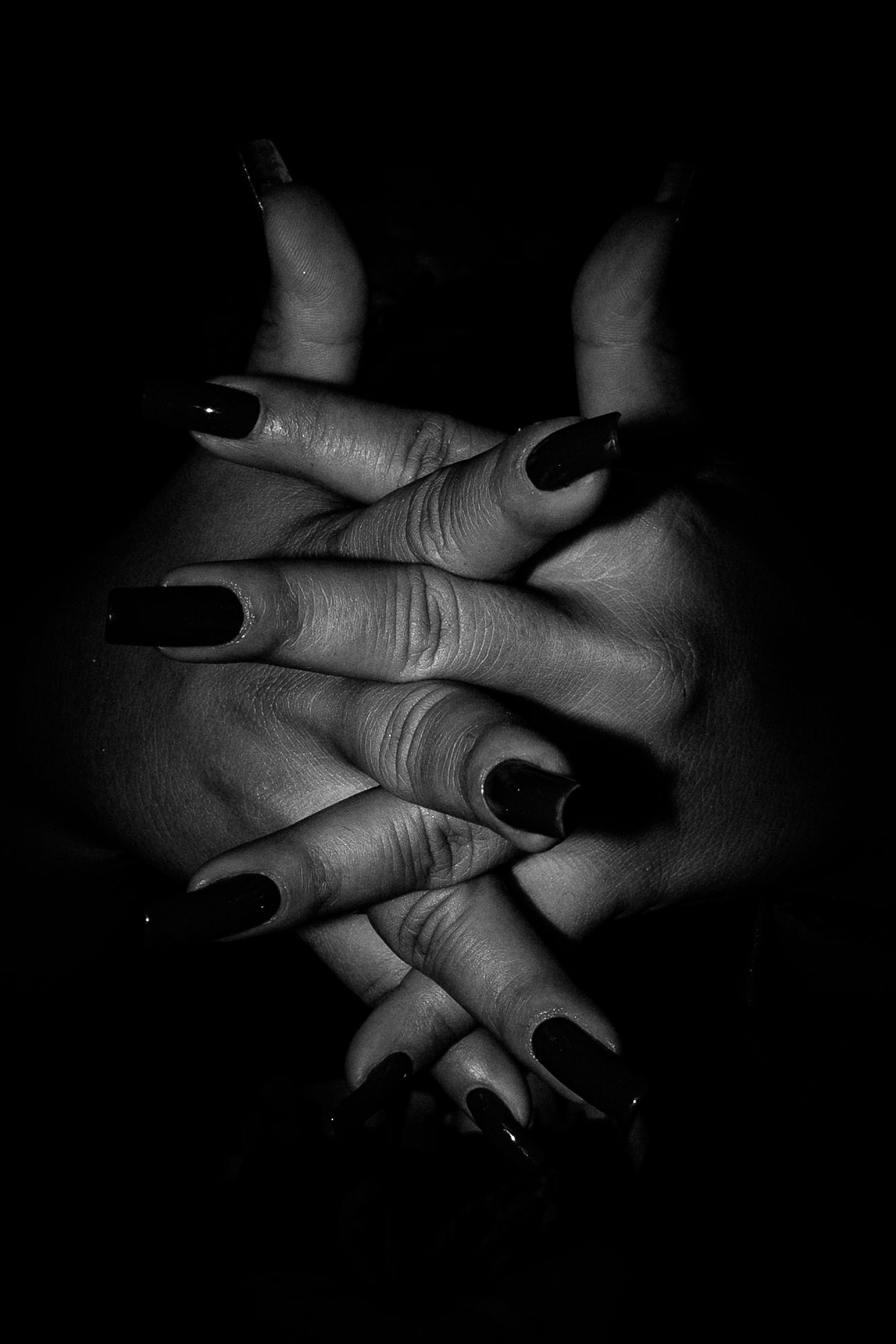 Manos entrelazadas con uñas negras, en blanco y negro sobre un fondo oscuro.